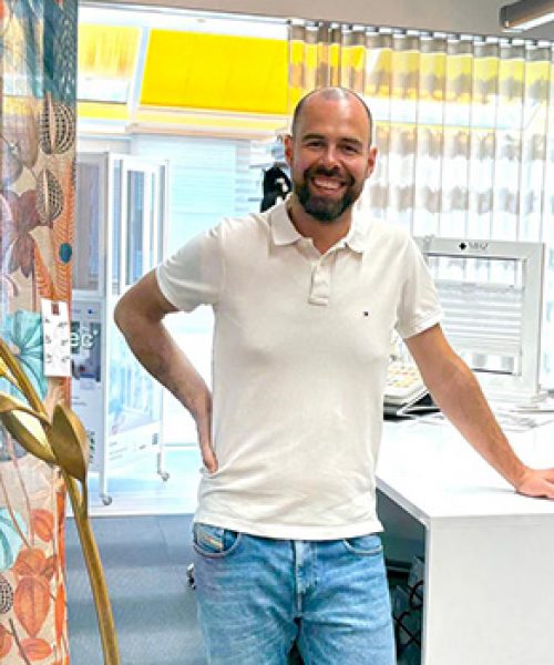 Inhaber Patrick Heckele im Laden an einem Tisch. Links eine Blumengardine und ein goldenes Dekoprodukt in Ährenform.
